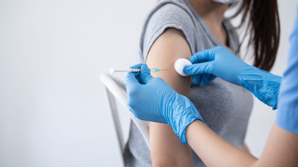 Woman receiving a vaccine injection in her upper arm at a clinic, administered by a healthcare professional wearing gloves.